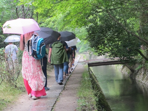 A group of students walking along the edge of a pond, using umbrellas for shade, at Tetsugaku-no-Michi, Kyoto, Japan 