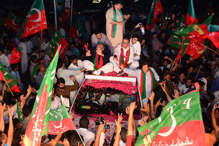 A car drives through a crowd of people waving the red and green flags of Pakistan's PTI political party. Then-PTI leader Imran Khan waves to the crowd from the car.