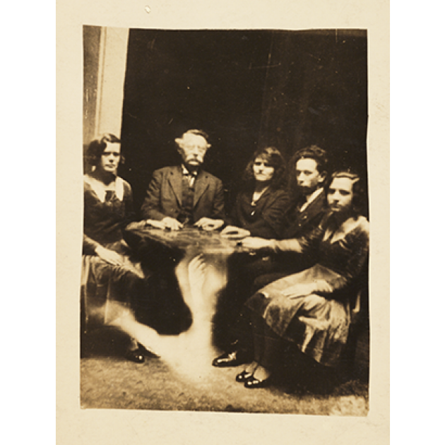 Black and white photograph of 2 men and 3 women around a table, each with one hand on top of it. A spectre of a hand arises from the floor toward the bottom of the table.