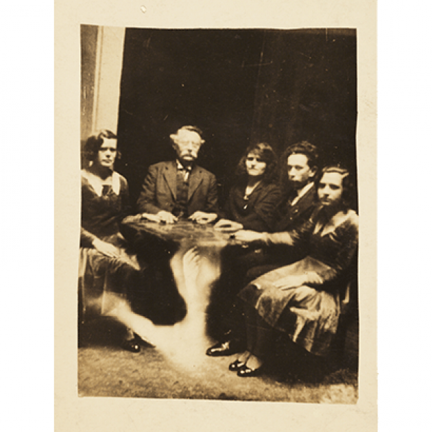  Black and white photograph of 2 men and 3 women around a table, each with one hand on top of it. A spectre of a hand arises from the floor toward the bottom of the table.