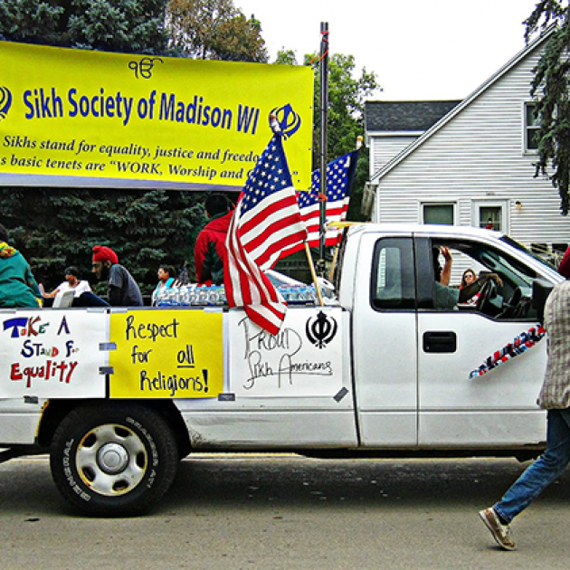 A white truck in a middle of a parade carries a yellow sign in its bed that reads "Sikh Society of Madison WI." Two American flags are on each side of the sign. A Sikh man wearing a dastar (turban) walks next to the truck and waves to the crowd  