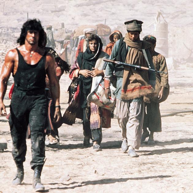 still from Rambo III. Sylvester Stallone in a black tank top leads a group of Afghan soldiers through a dusty landscape