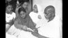 Gandhi seated, surrounded by people, writing a prayer message, January 18, 1948